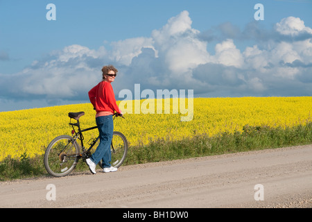 Femme à la mi-50's avec vélo sur route rurale à côté de champ en fleurs de canola, Manitoba, Canada Banque D'Images