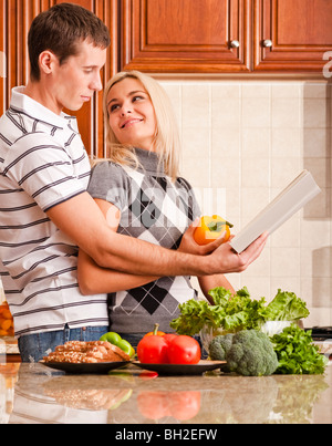 Jeune femme regarde en arrière à affectueusement un jeune homme pendant qu'il lit un livre de recettes. Un comptoir de cuisine est titulaire d'une variété de veget Banque D'Images