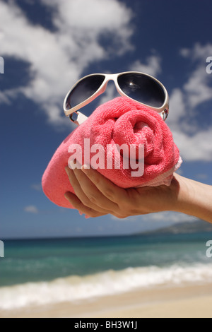 La main d'un homme tenant une serviette de plage rouge enroulé et une paire de lunettes blanche sur une plage tropicale Banque D'Images