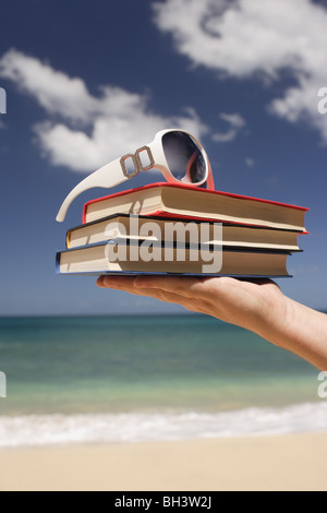 La main d'un homme tenant trois paperback books et une paire de lunettes blanche sur une plage tropicale Banque D'Images