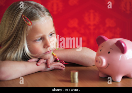 Young girl looking at a pink piggy bank et un tas de UK une livre de pièces sur une table, avec une expression grave Banque D'Images