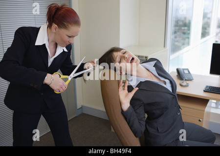 Une femme d'essayer de couper les cheveux d'une autre businesswoman assis à un bureau dans un bureau, avec une expression effrayée Banque D'Images
