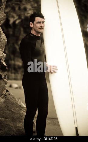 Un surfeur dans une combinaison isothermique holding his surfboard, debout sur la plage avec des rochers en arrière-plan sur l'océan. Banque D'Images