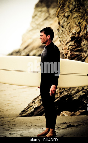 Un surfeur dans une combinaison isothermique holding his surfboard, debout sur la plage avec des rochers en arrière-plan sur l'océan. Banque D'Images