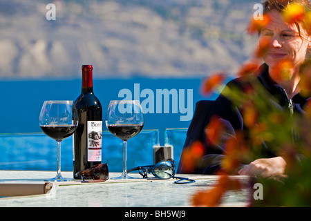 Femme assise à une table avec deux verres de vin rouge et une bouteille de vin avec vue sur le lac Okanagan à Bonitas Winery, Summerlan Banque D'Images