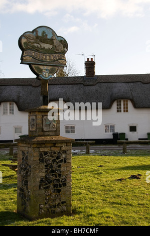 Le panneau du village de Weeting à Norfolk avec 'La ligne' cottages derrière. Banque D'Images