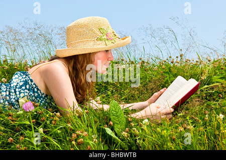 Young Woman Reading book in summer meadow with straw hat Banque D'Images