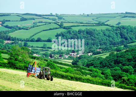 Agriculteur sur le tracteur d'ensilage d'herbe en coupe de foin ferme champ au-dessus de village de Winsford Somerset, England, UK. L'été. Banque D'Images