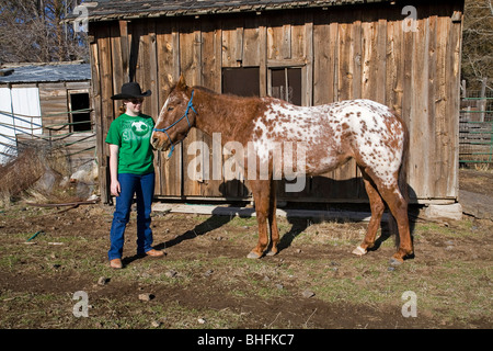 Un jeune cowgirl portant un chapeau de dix gallons et son cheval Appaloosa roan en face d'une ancienne grange. Banque D'Images