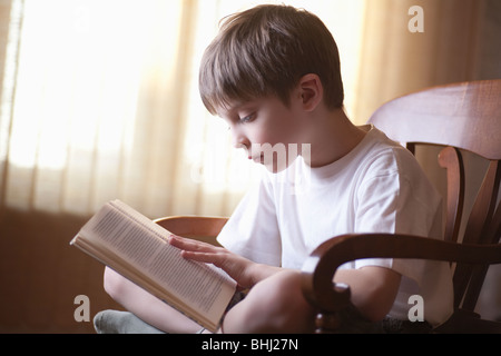 Garçon est assis en tailleur sur une chaise reading book Banque D'Images