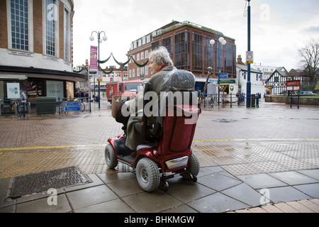 Une vieille dame dans un buggy électrique attend en bordure de la rue avant de traverser la rue sous la pluie, à Kingston, Surrey, Angleterre. Banque D'Images