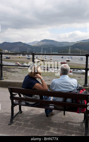 Couple d'âge moyen de vacanciers assis sur banc quai avec vue sur bateaux dans un port à marée basse Banque D'Images