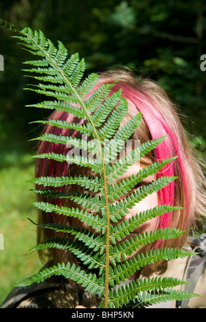 Une jeune fille aux cheveux teints en rose bob se cache derrière une feuille de fougère d'un vert vif qui masque partiellement son visage Banque D'Images