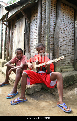 Les garçons s'asseoir dans un village traditionnel de la lecture d'un sol en bois guitare, Madagascar, océan indien, afrique Banque D'Images