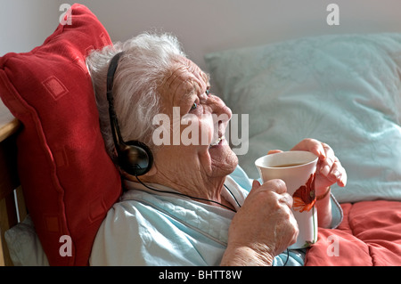 PERSONNE ÂGÉE DAME SOIN SOIGNANT contenu heureux femme âgée au lit portant des écouteurs apprécie une tasse de thé dans sa chambre confortable et ensoleillée Banque D'Images
