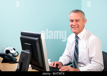 Mature businessman assis à un bureau Bureau de travailler sur son ordinateur souriant pour appareil photo. Banque D'Images