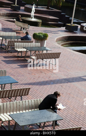 Quelqu'un lisant un livre sur les sièges et les tables à l'extérieur d'un café au bord de l'eau dans le Barbican London UK Banque D'Images