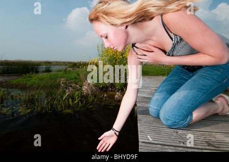 Jeune fille blonde assise sur un bord de marche touchent l'eau et à la recherche dans le noir profond de l'eau. Banque D'Images