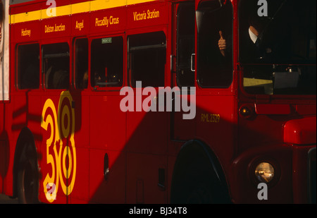 Conducteur d'un certain nombre 38 London bus rouge qui passe entre soleil et ombre, donne un coup de pouce à un autre conducteur. Banque D'Images