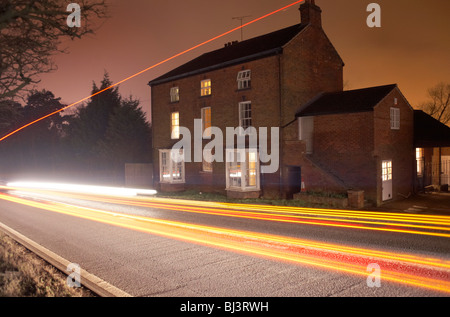 Se déplacer rapidement au-delà d'un bâtiment de ferme sur une longue UK UNE ROUTE, trafic invisible laisse sa légèreté sur une sombre nuit. Banque D'Images