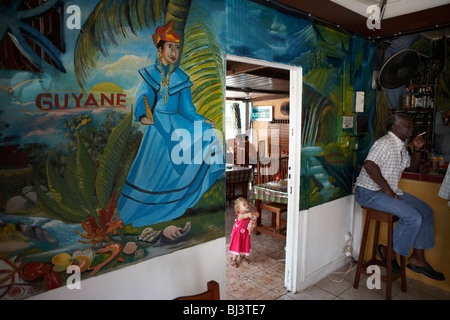Tenant sa poupée, un jeune enfant blanc portant une robe rose explore le Délice Restaurant à vieux Kourou, Guyane française. Banque D'Images