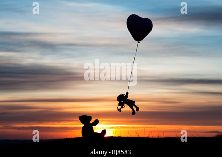 Poupée de chiffon holding sur un ballon d'hélium en forme de coeur s'envoler d'un ours au lever du soleil. UK. Still Life silhouette Banque D'Images