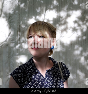 Portrait de jeune femme blonde avec frange et dress posing à motifs blanc en face de mur recouvert de feuilles d'ombre Banque D'Images