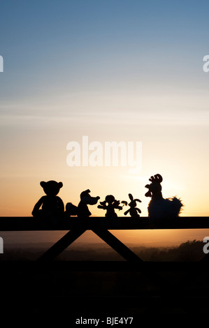 Poupée de chiffon, poulet, lapin, renard et l'ours peluches assis sur une barrière au coucher du soleil dans la campagne anglaise. Silhouette Banque D'Images