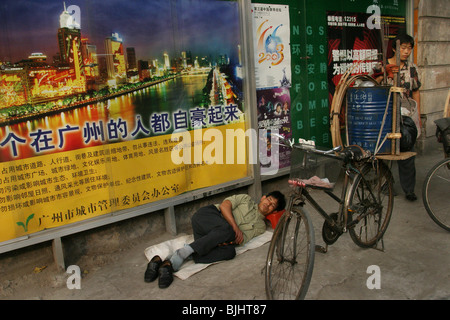 Un travailleur dort dans la rue ci-dessous une affiche montrant une nuit coup de la rivière des Perles de Guangzhou, Chine. Banque D'Images