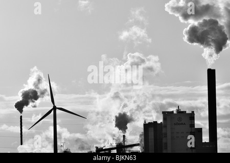 Image noir et blanc des volutes de fumée dans le ciel d'une usine comme cheminées du une éolienne se trouve dans l'avant-plan Banque D'Images