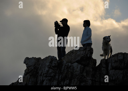 Silhouette d'un homme prenant photo d'un paysage debout sur un rocher avec femme et chien, au coucher du soleil, Hermanus, South Western Ca Banque D'Images