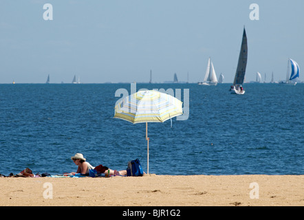 Femme en train de bronzer SUR LA PLAGE DE BRIGHTON AVEC PARE-SOLEIL PARASOL ET BATEAUX SUR PORT PHILLIP BAY VICTORIA Melbourne Australie Banque D'Images