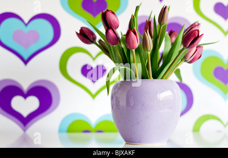 Un vase de tulipes violet en face de papier peint couleur décoré d'un motif coeur Banque D'Images