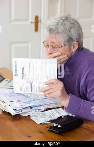 Choqué senior woman retraité pensionné avec un gros tas de billets avec les main sur sa bouche l'air inquiet à un grand relevé de carte de crédit. UK Banque D'Images