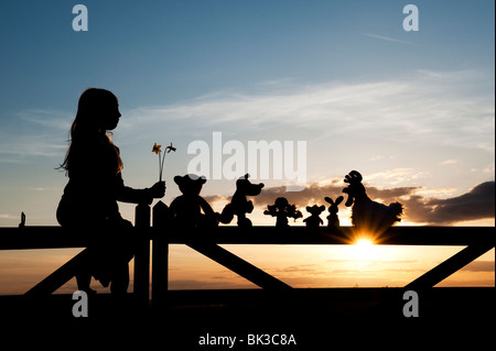 Jeune Fille de jonquilles et une poupée de chiffon, poulet, lapin, renard et l'ours peluches assis sur une barrière au coucher du soleil . Silhouette Banque D'Images