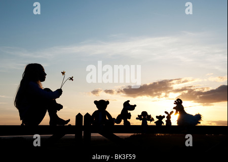 Jeune Fille de jonquilles et une poupée de chiffon, poulet, lapin, renard et l'ours peluches assis sur une barrière au coucher du soleil . Silhouette Banque D'Images