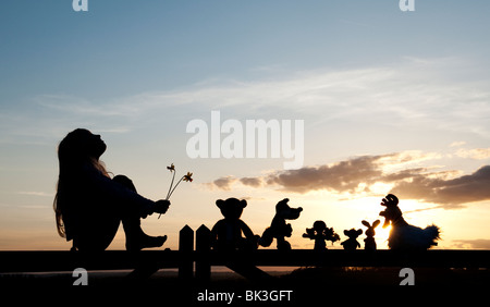 Jeune Fille de jonquilles et une poupée de chiffon, poulet, lapin, renard et l'ours peluches assis sur une barrière au coucher du soleil . Silhouette Banque D'Images