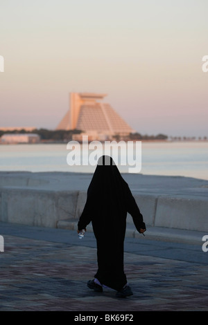 Femme arabe portant une robe noire traditionnelle balade le long de la Corniche, l'hôtel Sheraton à l'arrière, à Doha, Qatar Banque D'Images