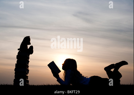 Jeune fille couchée la lecture d'un livre en face d'un ours assis sur une pile de livres au coucher du soleil. Silhouette Banque D'Images