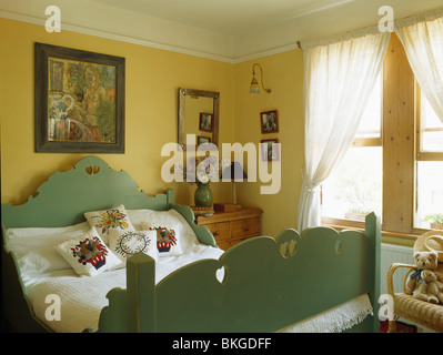 Photo sur le mur au-dessus de lit en bois peint en vert avec coussins brodés en jaune chambre avec des rideaux de voile blanc sur la fenêtre Banque D'Images