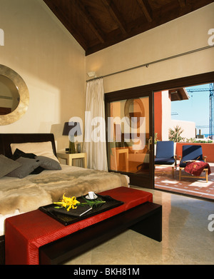 Tabouret rouge ci-dessous lit avec fausse fourrure plaids en espagnol moderne chambre avec portes-fenêtres donnant sur un balcon Banque D'Images