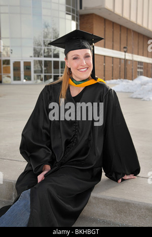 Attractive blonde woman in graduation uniforme assis en face de college building and smiling Banque D'Images