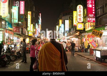 Les gens passent devant un moine, crowded street scène montrant les panneaux d'éclairage sans fin et les gens la nuit shopping dans la rue du Marché de nuit de Huaxi Touristiques Banque D'Images