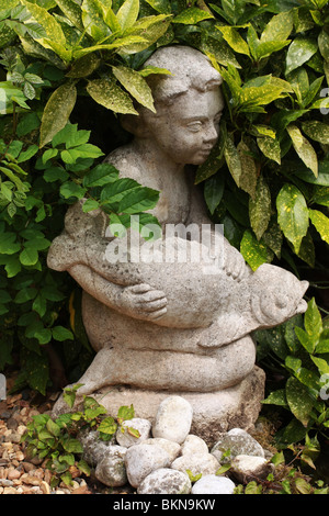 Beau jardin statue en pierre, d'un enfant et le poisson entourée de feuilles vertes, entouré de pierres rondes et de gravier. Banque D'Images