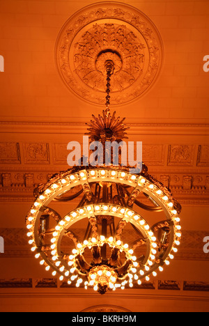 Lustres du Grand Central Station Vanderbilt Hall New York City | NEW YORK, New York — regardant droit devant un lustre doré et son médaillon de plafond assorti dans le Vanderbilt Hall du Grand Central terminal. La rosette rayonnante ci-dessus dissimule le point de suspension, dispositif décoratif commun aux intérieurs Beaux-Arts du début du XXe siècle. Des rangées de petites ampoules sonnent à chaque niveau, produisant la lueur ambre caractéristique de la salle. Banque D'Images