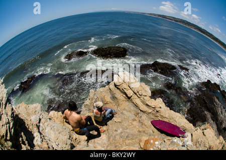 Un jeune homme et femme assis sur une pointe rocheuse tout en regardant les surfeurs à Punta de Lobos, Puchilemu, au Chili. (Fisheye) Banque D'Images