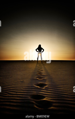 Jeune femme en distance standing on beach at sunset avec empreintes de pieds dans le sable Banque D'Images