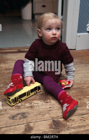ENFANT EN BAS âge, JOUER, PLANCHER : un enfant de deux ans bébé fille enfant en bas âge jouant avec un bus jouet avec une boîte de bonbons en chaussettes et bottes modèle libéré Banque D'Images