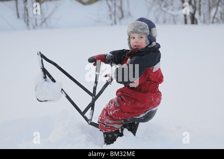 GARÇON TOUT-PETIT ASSIS SUR LA GRUE JOUET DANS LA NEIGE PROFONDE DE L'HIVER : un enfant de bébé garçon de deux ans joue avec la pelleteuse dans le modèle de parc de neige profond libéré Banque D'Images