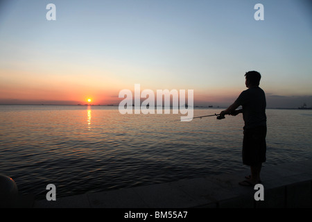 Un pêcheur jette sa ligne dans la baie de Manille à partir de Roxas Boulevard au coucher du soleil près de la zone en malate de Manille, Philippines. Banque D'Images